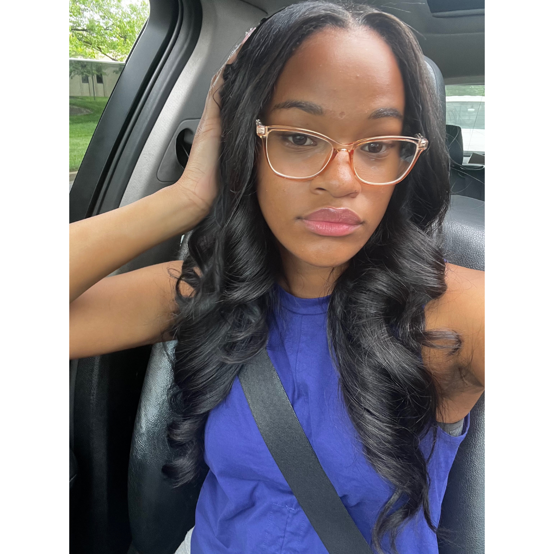 Woman sitting in a car wearing glasses and a blue shirt. wearing raw loose wave elite glam society bundles